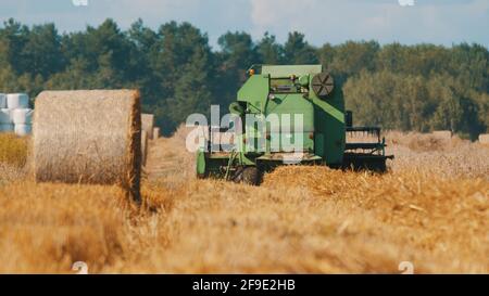 Warsaw, Poland 10.08.2020 - Backview of a big green tractor in an agricultural field. Hay stacks can be seen on the left side. A golden wheat field during the daytime with trees against blue sky Stock Photo