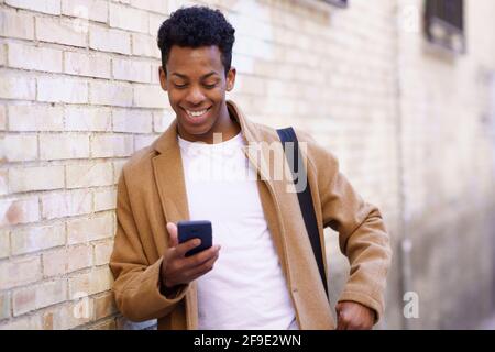 Young black man typing on his smartphone leaning against a brick wall in the street. Stock Photo