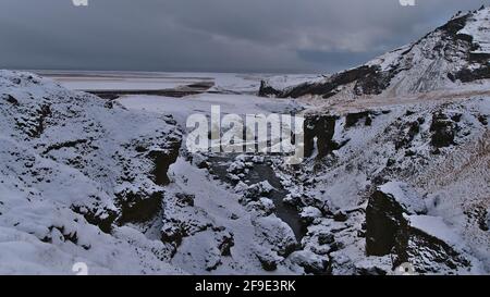 View of Skógá river flowing through rocky canyon on the southern coast of Iceland in winter season with snow-covered rugged mountains. Stock Photo