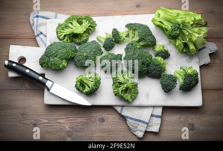 Sicilian broccoli cut into pieces with white marble cutting board and knife on wooden background, top view. Stock Photo