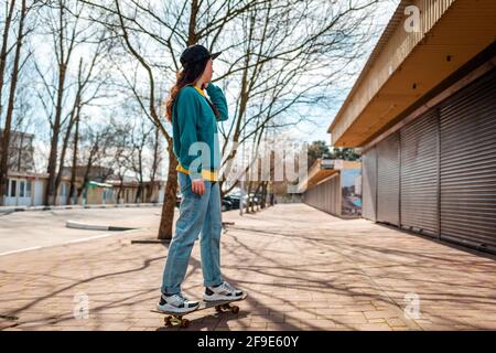 A young Caucasian hipster woman rides down the street on a skateboard. In the background, an alley. Copy space. Concept of sports lifestyle and street Stock Photo