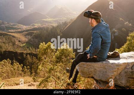 Side view of male traveler in VR glasses interacting with virtual reality while sitting on hill in mountainous terrain at sunset Stock Photo