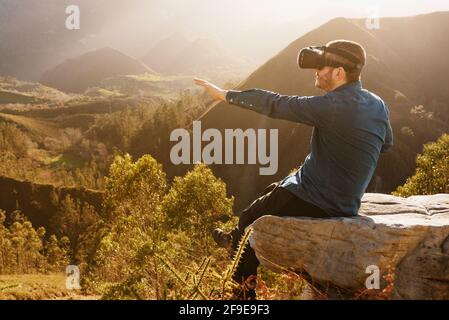 Side view of male traveler in VR glasses interacting with virtual reality while sitting on hill in mountainous terrain at sunset Stock Photo