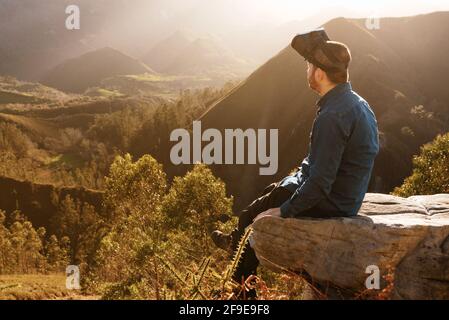 Side view of male traveler in VR glasses interacting with virtual reality while sitting on hill in mountainous terrain at sunset Stock Photo