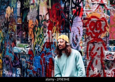 Dreamy hipster young male looking away while standing against graffiti wall in city Stock Photo