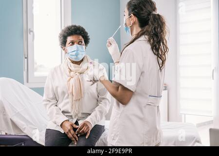 Female medical specialist in protective uniform, latex gloves and face mask doing nasal coronavirus test on African American mature Woman patient in c Stock Photo
