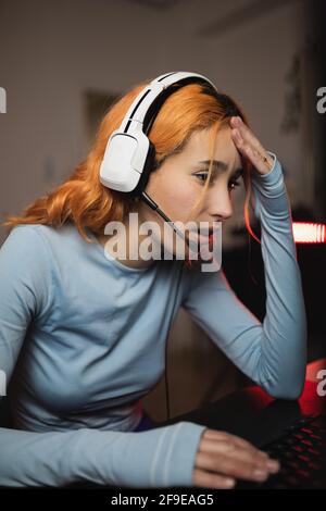 Side view of worried female gamer in headphones playing video game while sitting at table with keyboard Stock Photo
