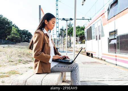 Side view of focused Asian female in mask sitting on bench at railway station and browsing laptop while waiting for train Stock Photo