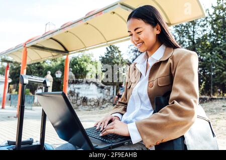 Asian female in mask sitting on bench at railway station and browsing laptop while waiting for train Stock Photo