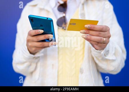 Cropped unrecognizable stylish African American female paying with plastic card during online shopping via mobile phone while standing on blue backgro Stock Photo