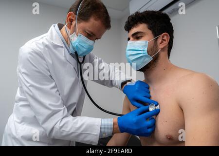 Low angle of male physician in mask using phonendoscope while examining lunges of patient in mask in hospital Stock Photo