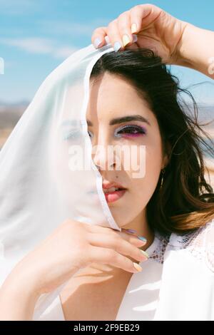 Young brunette Woman covering eye with veil while looking at camera on wedding day Stock Photo