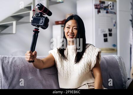Smiling ethnic female vlogger recording video on photo camera while sitting on couch in living room Stock Photo