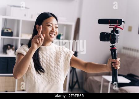 Smiling ethnic female vlogger recording video on photo camera while making hand gestures standing in living room Stock Photo