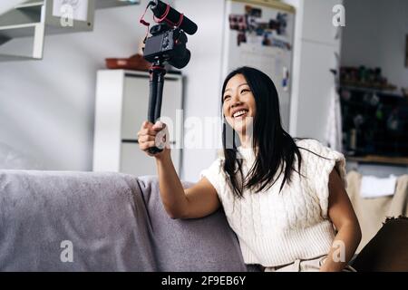 Smiling ethnic female vlogger recording video on photo camera while sitting on couch in living room Stock Photo