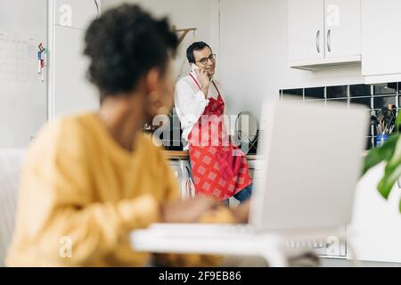 man in apron speaking on smartphone in kitchen and black Woman browsing laptop at home Stock Photo