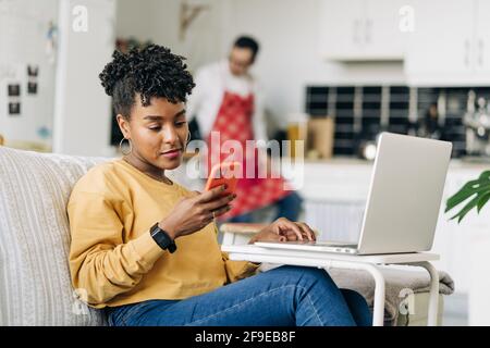 man in apron browsing smartphone in kitchen and black Woman browsing laptop and smartphone at home Stock Photo