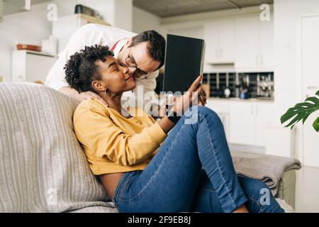 Side view of tender man kissing black Woman sitting on sofa and reading book at weekend Stock Photo