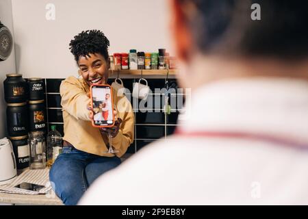 Cheerful African American female showing taken photo on smartphone for unrecognizable boyfriend while having fun in kitchen at home Stock Photo