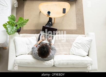 Young hipster male with curly hair browsing internet on netbook while resting on couch in house room Stock Photo
