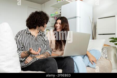 Cheerful Woman with netbook talking to tattooed boyfriend while looking at each other on couch in house room Stock Photo