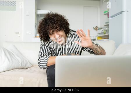 Content male with tattoos showing hi gesture against netbook during video chat while sitting on couch in apartment Stock Photo