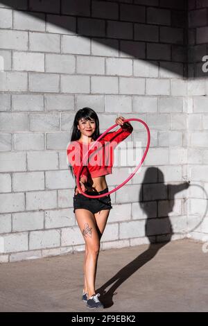 Young tattooed Woman in activewear twirling hula hoop while dancing against brick walls with shadows Stock Photo