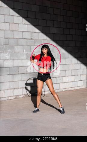 Young tattooed Woman in activewear twirling hula hoop while dancing against brick walls with shadows Stock Photo