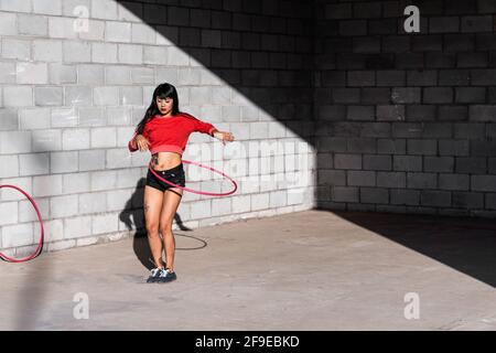 Young tattooed Woman in activewear twirling hula hoop while dancing against brick walls with shadows Stock Photo