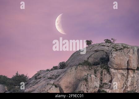 Low angle of spectacular scenery of rough rocky mountain under pink sunset sky with waning moon in Sierra de Guadarrama National Park Stock Photo