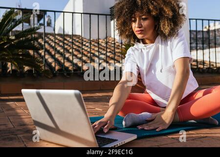 Focused African American female with curly hair choosing online tutorial on laptop while sitting on mat on rooftop and preparing for yoga lesson Stock Photo
