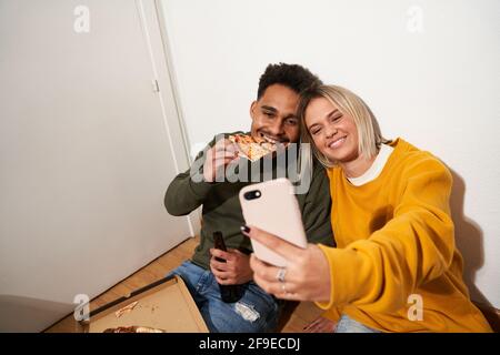 From above of positive multiracial couple eating tasty pizza and taking self shot on smartphone while chilling together at home Stock Photo