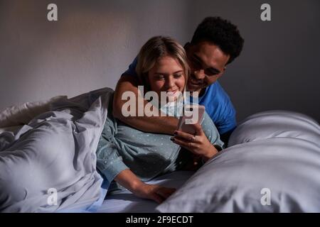 Delighted multiracial couple lying on bed while browsing smartphone and having fun together in morning Stock Photo