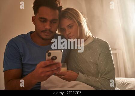 Calm multiracial couple hugging in morning while sitting on bed and using smartphone together Stock Photo