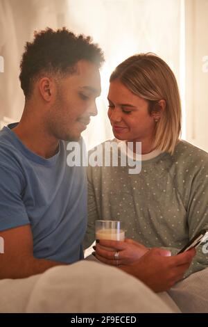 Delicate multiracial couple sitting on bed and enjoying morning together while drinking milk and using smartphone Stock Photo