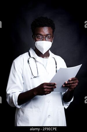 African American male doctor in protective mask and white uniform taking notes on clipboard while looking at camera on black background Stock Photo