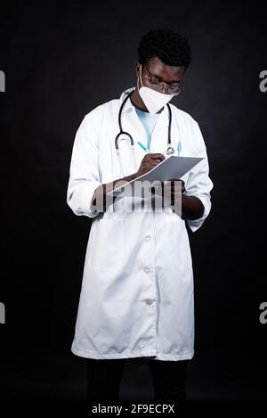 African American male doctor in protective mask and white uniform taking notes on clipboard while on black background Stock Photo