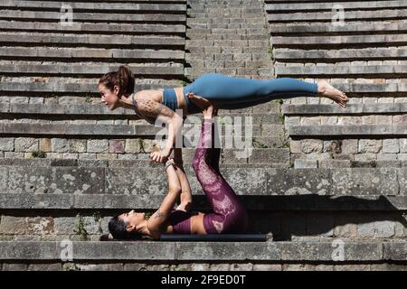 Side view of flexible female supporting Woman on legs while practicing acro yoga together on stone stairs Stock Photo