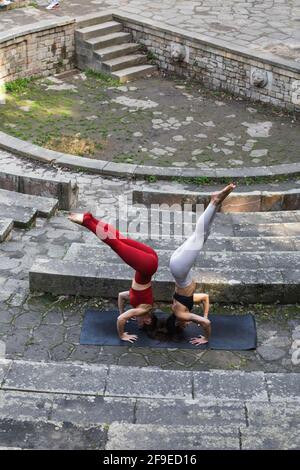 From above of fit females balancing in Salamba Sirsasana while practicing yoga together on mat on stone stairs Stock Photo