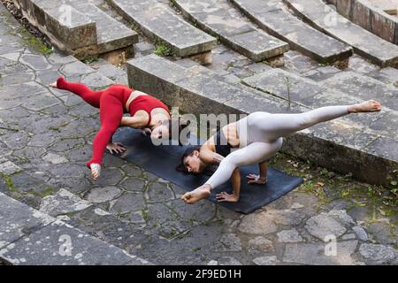 High angle of concentrated flexible females in activewear balancing in Eka Pada Koundinyasana on mat while practicing yoga on stone stairs Stock Photo