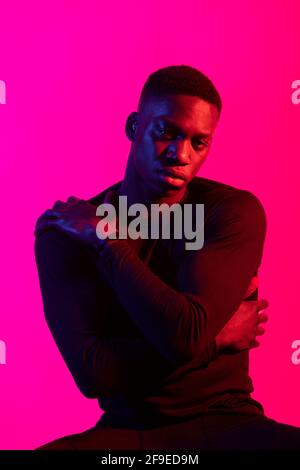 Confident young black man in dark sportive outfit looking at camera on neon pink background in dark studio Stock Photo