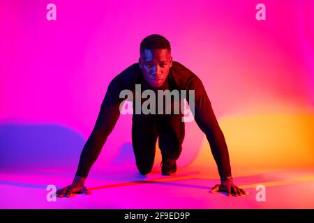 Full length concentrated fit African American male runner standing in crouch start position and looking at camera on colorful background in studio Stock Photo