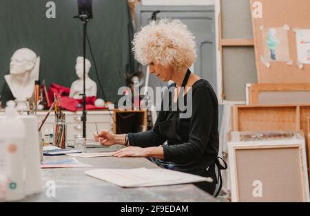 Focused female artist sitting at table and painting with watercolors on paper while working in creative workshop Stock Photo
