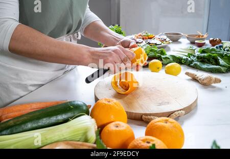 Crop anonymous female peeling fresh orange with knife over cutting board at table with vegetables and fruits Stock Photo