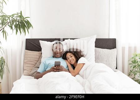 Smiling attractive millennial couple using smartphone, lying on bed together Stock Photo