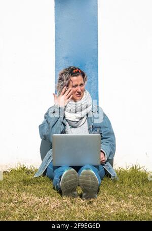A Caucasian woman sitting on the grass and leaning on a wall while working on her laptop in a park in Spain Stock Photo