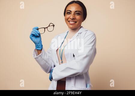 Positive woman doctor on brown background. Woman doctor holding her eyeglasses and smiling at camera. Stock Photo