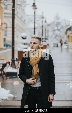 A handsome young confident man in a black winter outfit with a brown scarf strolling around the city Stock Photo