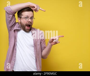 Funny man points index fingers at something, he stands in an unusual position and happily looks at the camera. Copy space.  Stock Photo