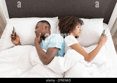 Awake using phone for chat, couple in quarrel, lying on bed back to back, using smartphones Stock Photo